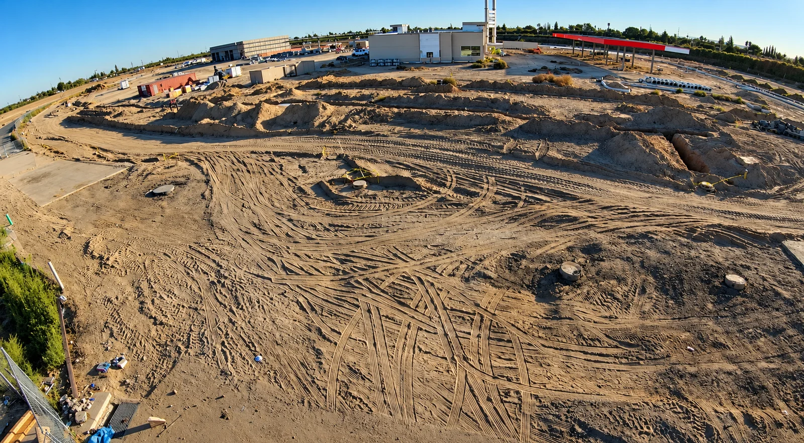 Construction site from another angle, showing earthmoving tracks, utility access points, trenches, and stacked pipe sections.