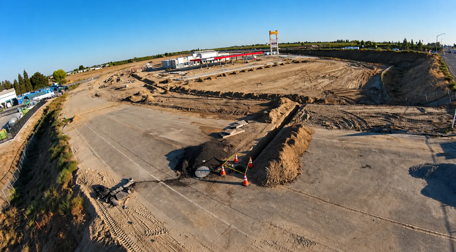Wide view of a construction site with excavated trenches, graded earth, and traffic cones around a utility access point.